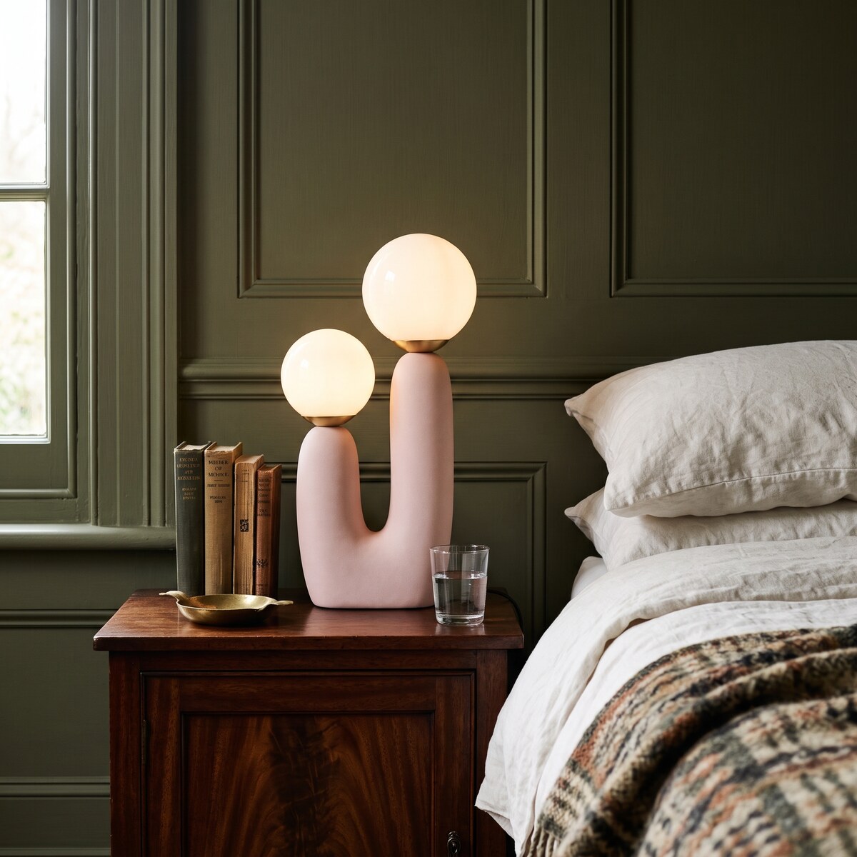 Cacti blush pink table lamp glowing on an antique bedside table against an olive-green panelled wall
