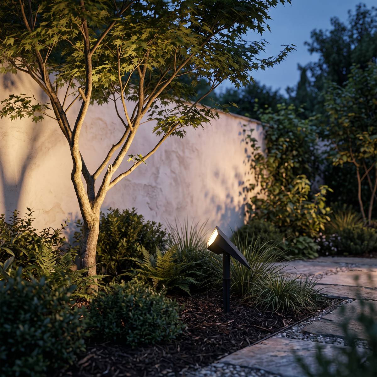 Sunpeak solar spotlight uplighting a small tree against a garden wall at dusk